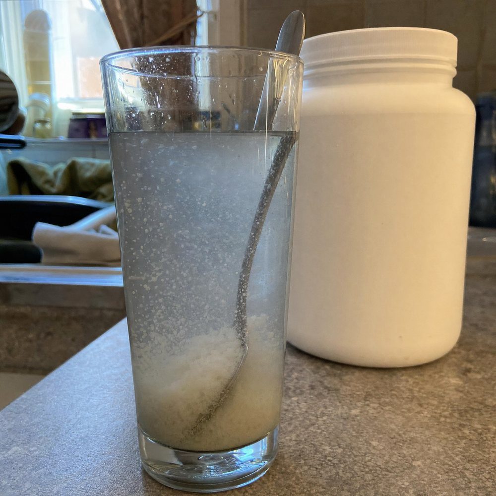 Ein Glas Wasser, in dem sich billiges Kreatin-Pulver nicht auflöst und wie Sand am Boden liegt, dahinter eine namenlose weiße Dose