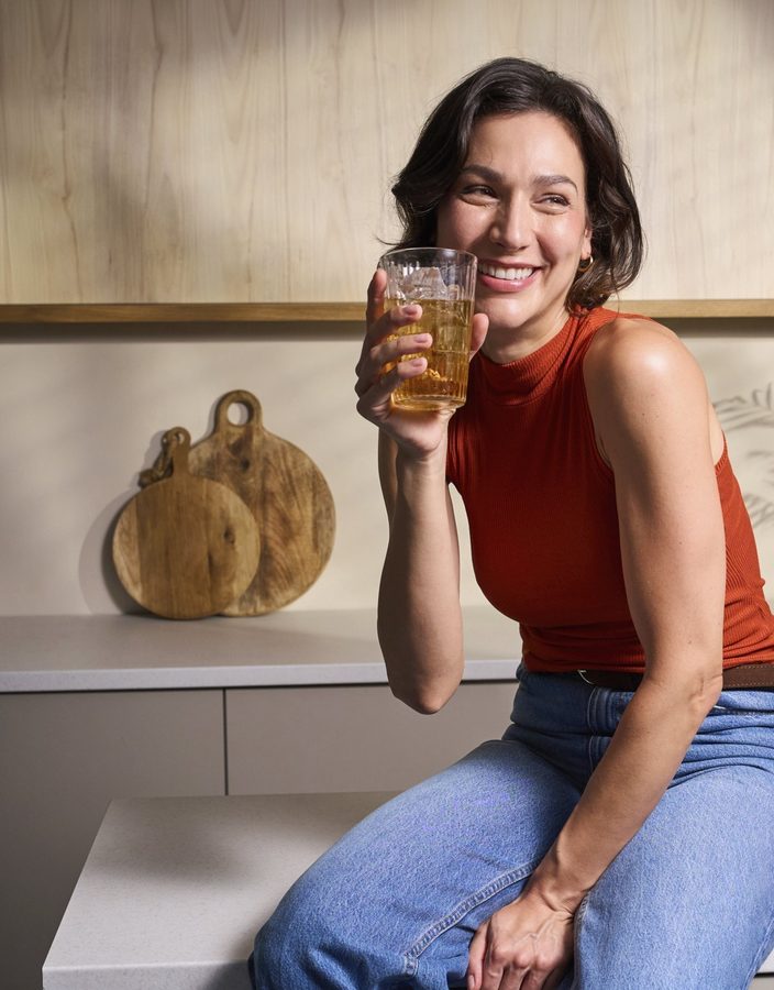Frau lacht morgens in ihrer Küche mit einem Glas in der Hand, voller Energie dem Bett auf