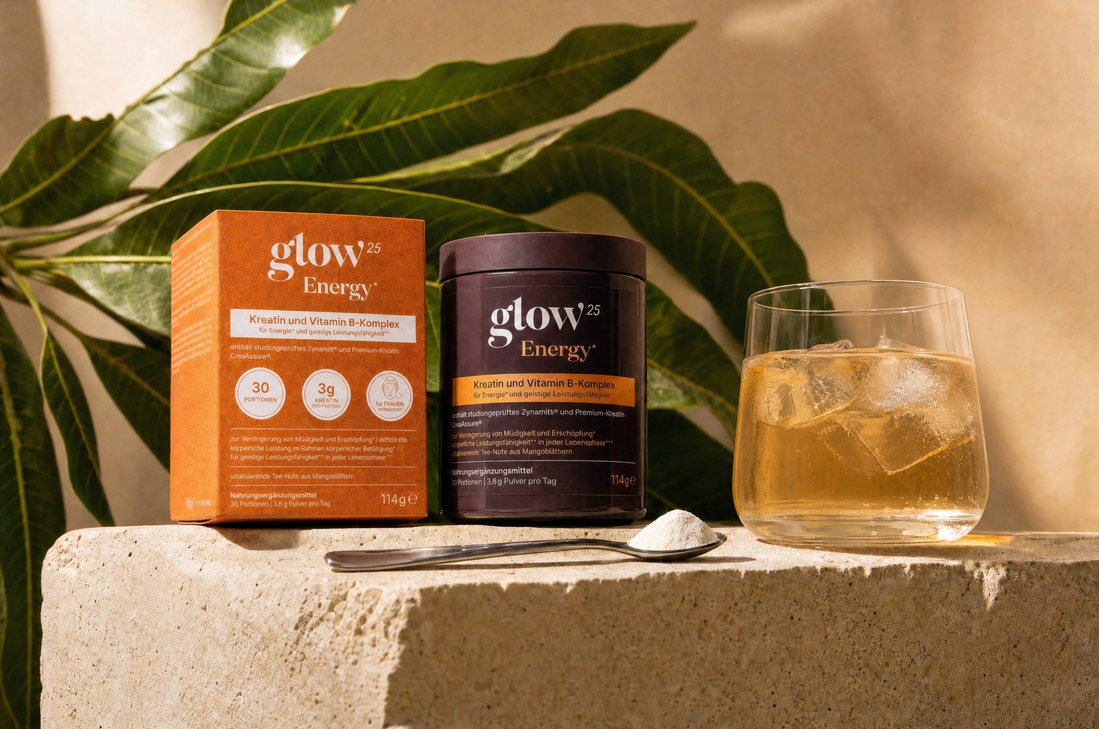 Glow25 Energy® Box und Dose auf Travertin, daneben Mangoblätter, Löffel und ein Glas mit Eis-Getränk