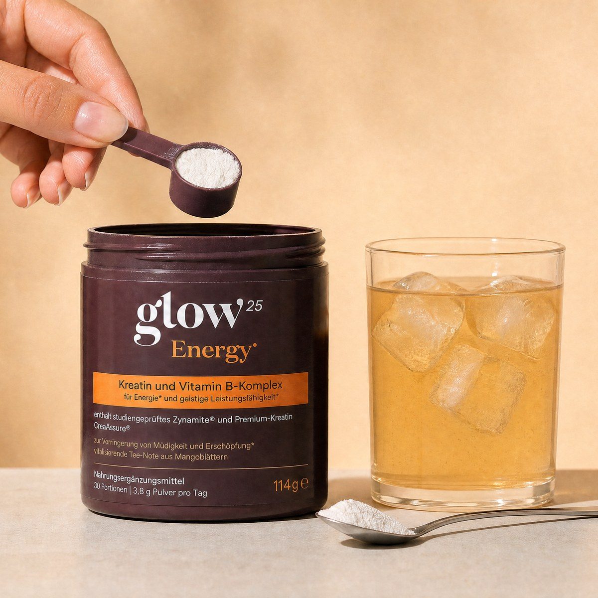 Eine Hand hebt den Dosierlöffel aus der Glow25 Energy Dose, daneben ein Löffel mit Pulver und ein Glas mit Eis-Getränk