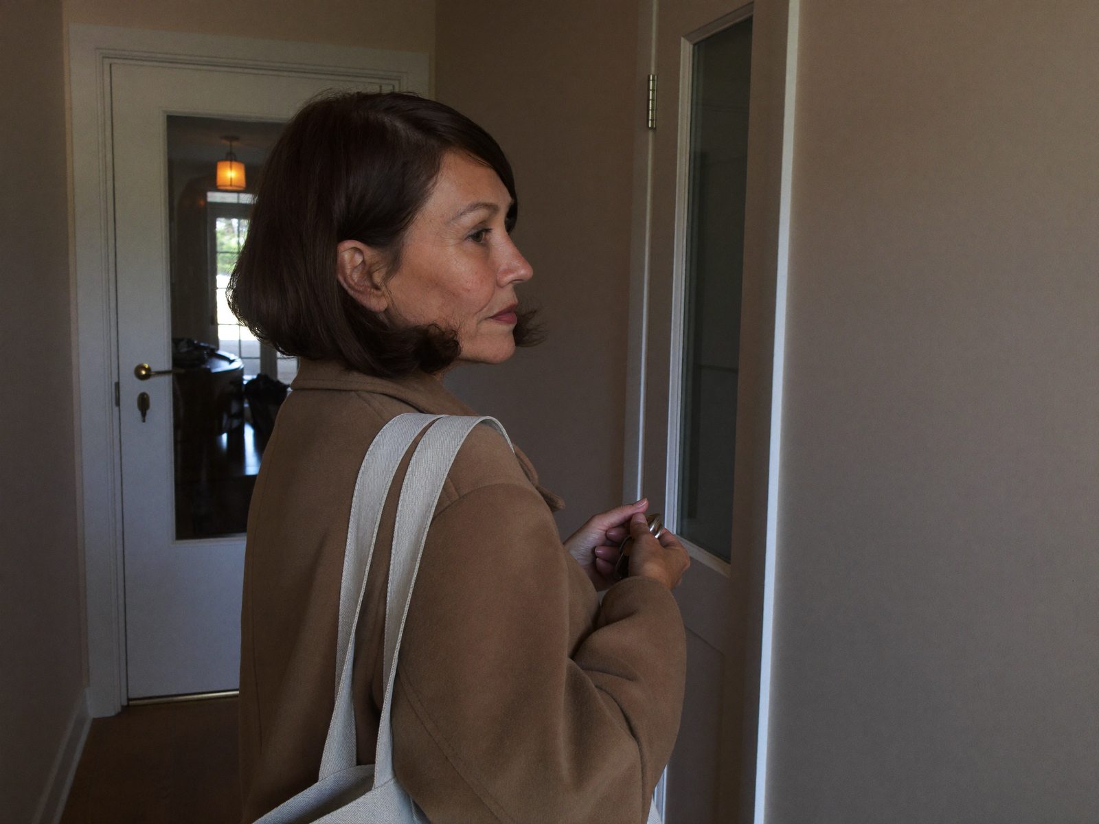 Femme en manteau dans l'entrée tôt le matin, le regard tourné vers la porte d'entrée, prête à partir